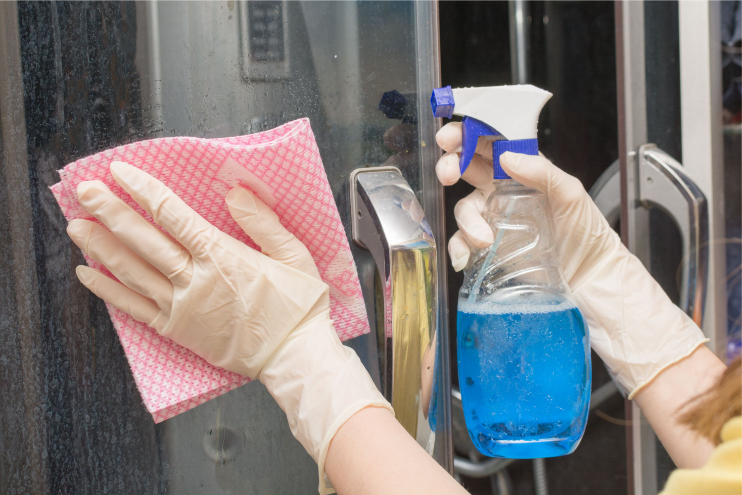 a close up of gloved hands holding a cloth and cleaning spray while cleaning a shower screen